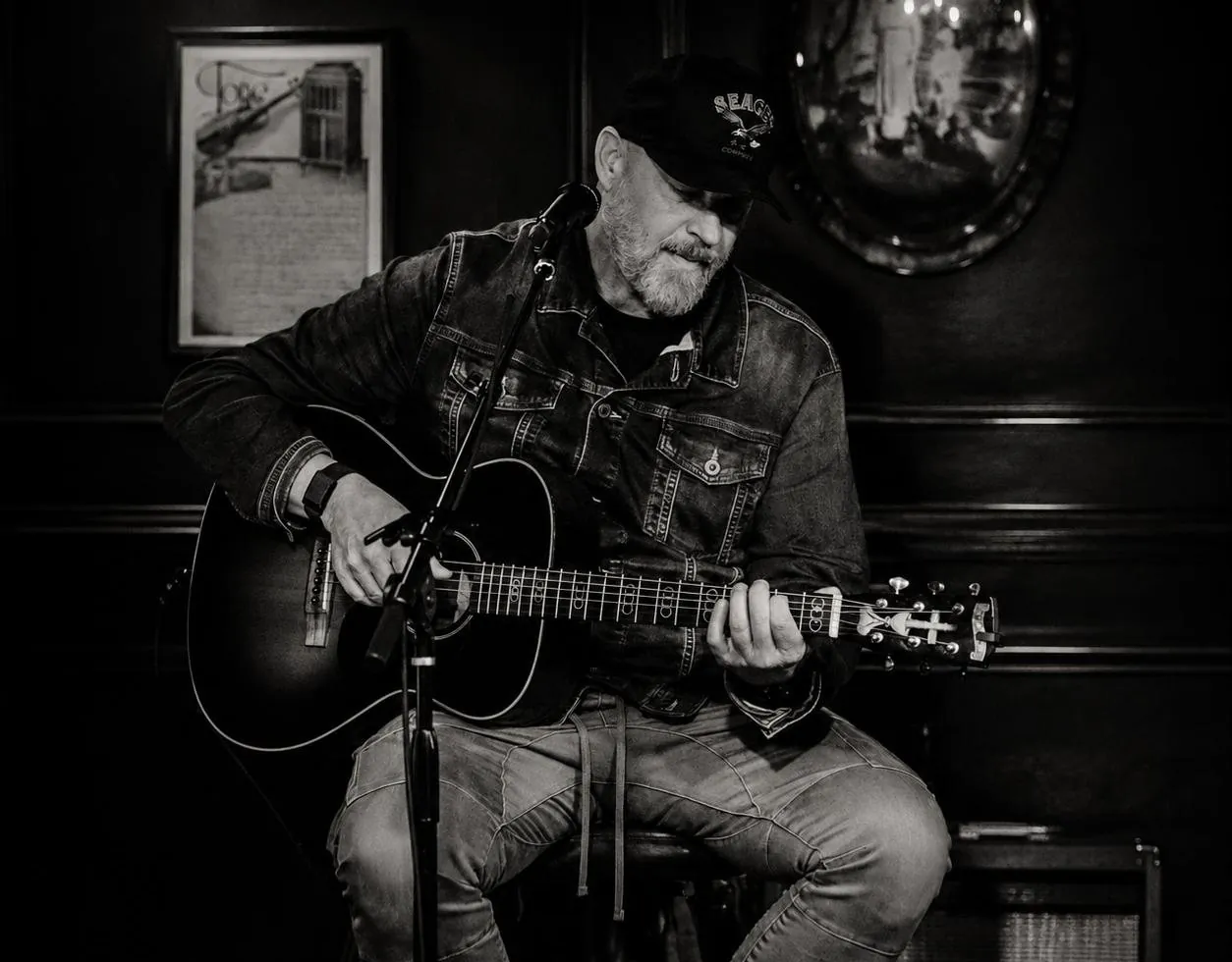 Anthony Skinner playing an acoustic guitar at a microphone in a dark wood-paneled room, in black and white