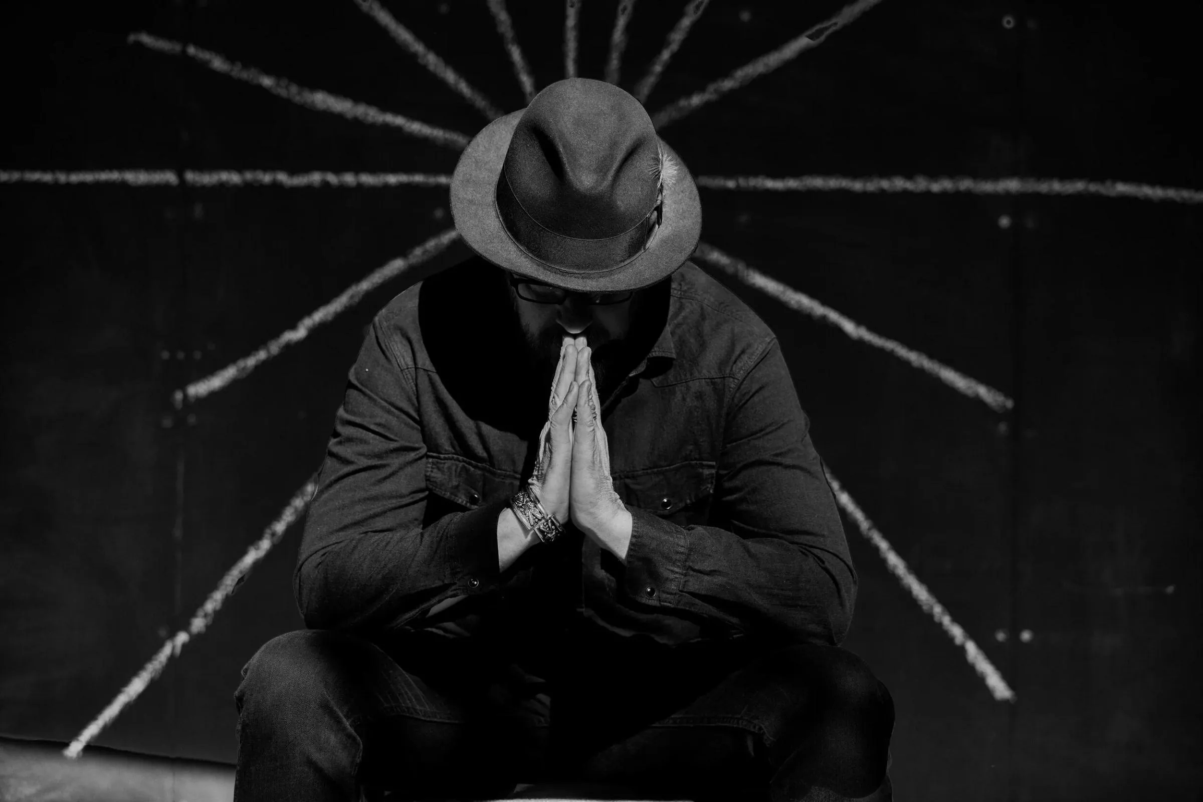 Anthony Skinner kneeling in prayer in a hat, hands pressed together, chalk lines radiating behind him, in black and white