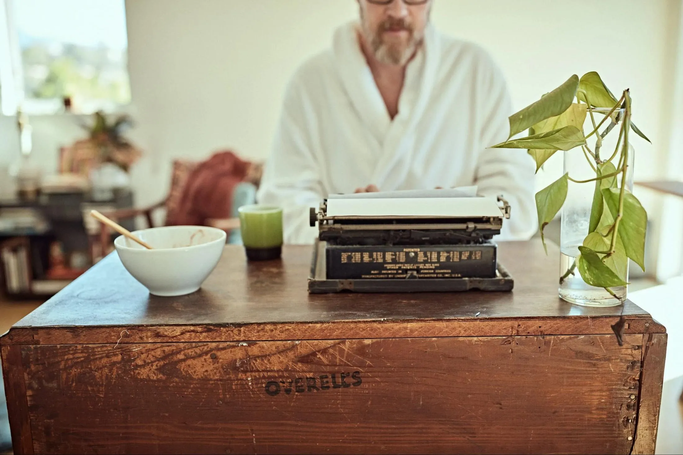 Anthony Skinner in a white robe typing on a vintage typewriter at a weathered wooden desk in morning light